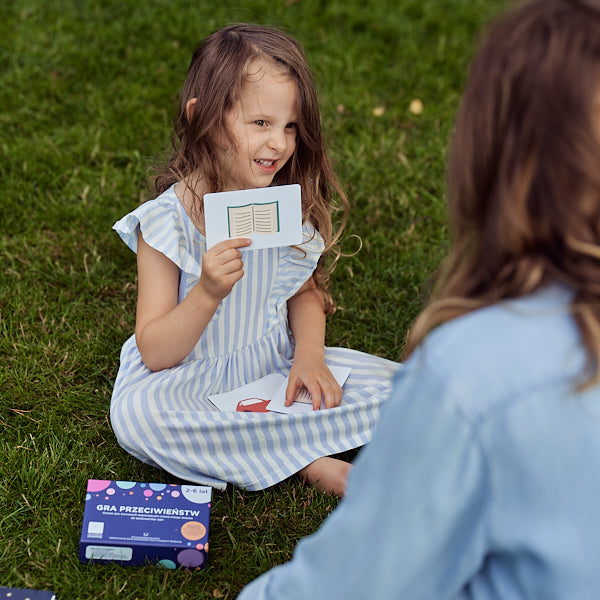 Obrazek przedstawia dziewczynkę prezentującą siedzącej naprzeciwko niej kobiecie jedną z kart "Gry przeciwieństw".