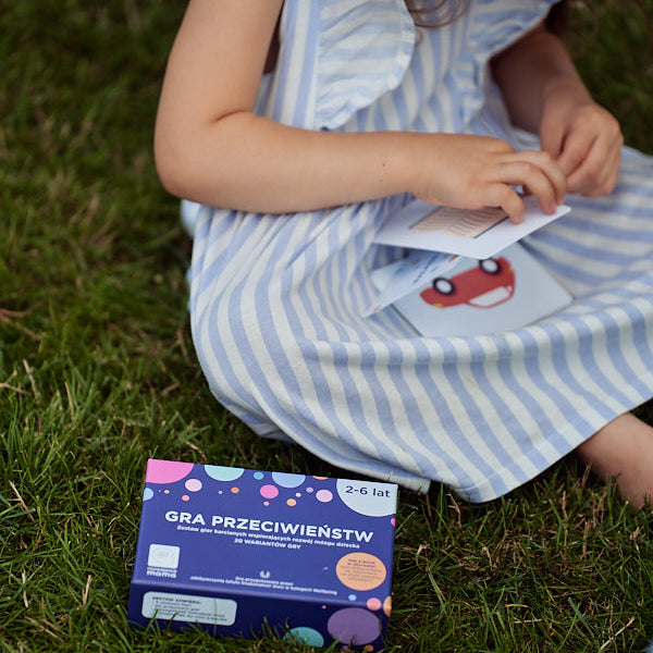 Obrazek przedstawia pudełko "Gry przeciwieństw", leżące na trawie. Obok siedzi dziewczynka i przegląda karty do gry.
