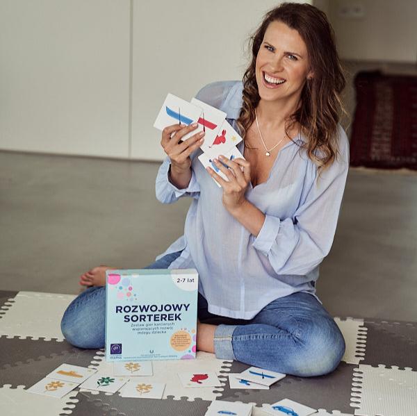 Zdjęcie przedstawia kobietę siedzącą na podłodze i prezentującą karty na gry "Rozwojowy sorterek". Obok widoczne pudełko gry.