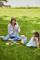 Zdjęcie przedstawia siedzącą na trawie nad rzeką kobietę, prezentującą jedną z kart aktywności odwróconej bokiem do obiektywu dziewczynce. Na trawie rozłożone pudełko gry "Kostka aktywności", kostka i karty z zadaniami.