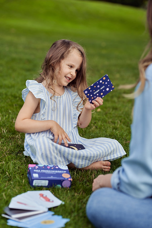 Obrazek przedstawia dziewczynkę siedzącą na trawie i grającą w "Grę przeciwieństw".