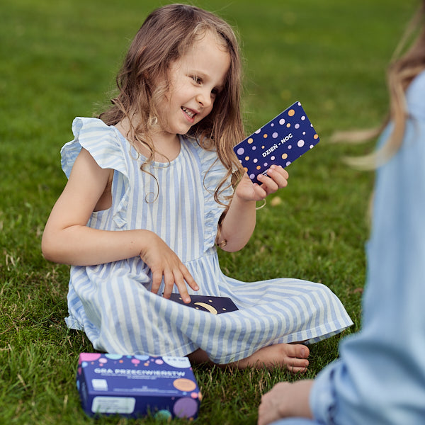 Obrazek przedstawia dziewczynkę siedzącą na trawie i grającą w "Grę przeciwieństw".