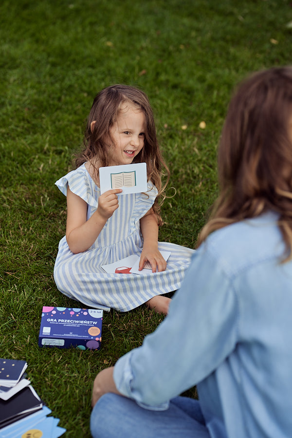 Obrazek przedstawia dziewczynkę prezentującą siedzącej naprzeciwko niej kobiecie jedną z kart "Gry przeciwieństw".