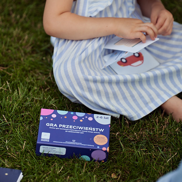 Obrazek przedstawia pudełko "Gry przeciwieństw", leżące na trawie. Obok siedzi dziewczynka i przegląda karty do gry.