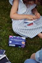 Obrazek przedstawia pudełko "Gry przeciwieństw", leżące na trawie. Obok siedzi dziewczynka i przegląda karty do gry.