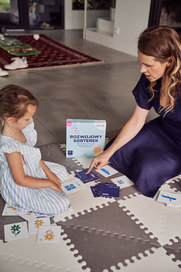 Zdjęcie przedstawia kobietę i dziewczynkę siedzące na podłodze i grające w grę "Rozwojowy sorterek".