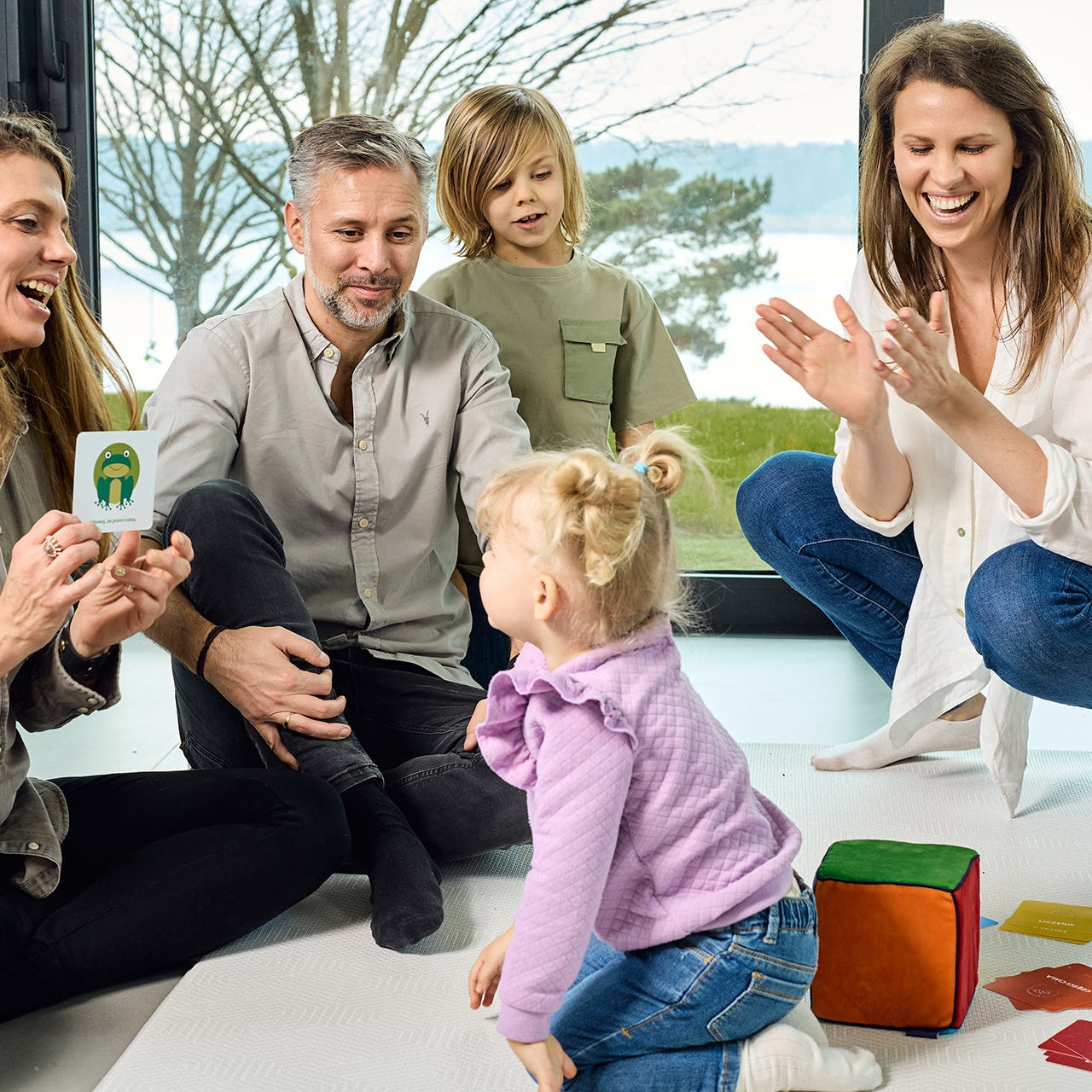 Rodzic i dziecko bawiący się Kostką Aktywności na dywanie. Dziecko rzuca kostką, a rodzic wyciąga kartę z zadaniem, co sprzyja budowaniu więzi i radosnej nauce.