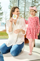 Na zdjęciu widoczna siedząca na podłodze kobieta prezentująca jedną z kart do gry "Kostka aktywności - nauka przez zabawę". Obok niej, opierając dłoń na jej ramieniu, stoi dziewczynka 