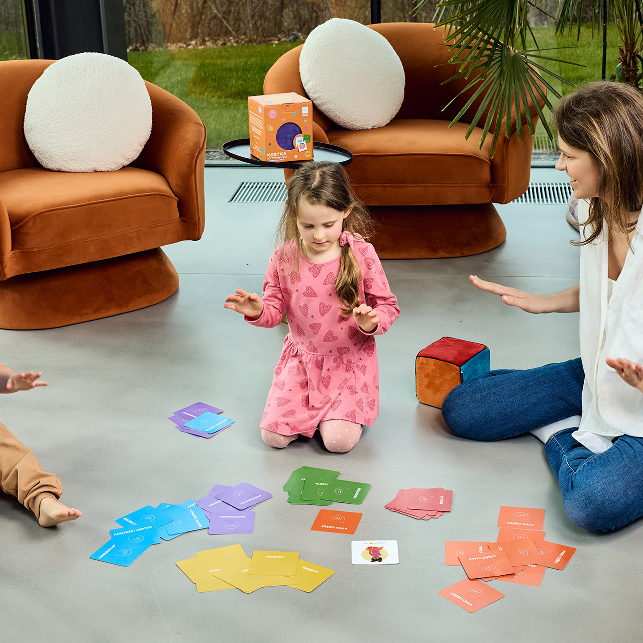 Na zdjęciu widocznych dwoje dzieci - chłopiec i dziewczynka - oraz kobieta. Wszyscy siedzą na podłodze i grają w grę "Kostka aktywności - nauka przez zabawę"