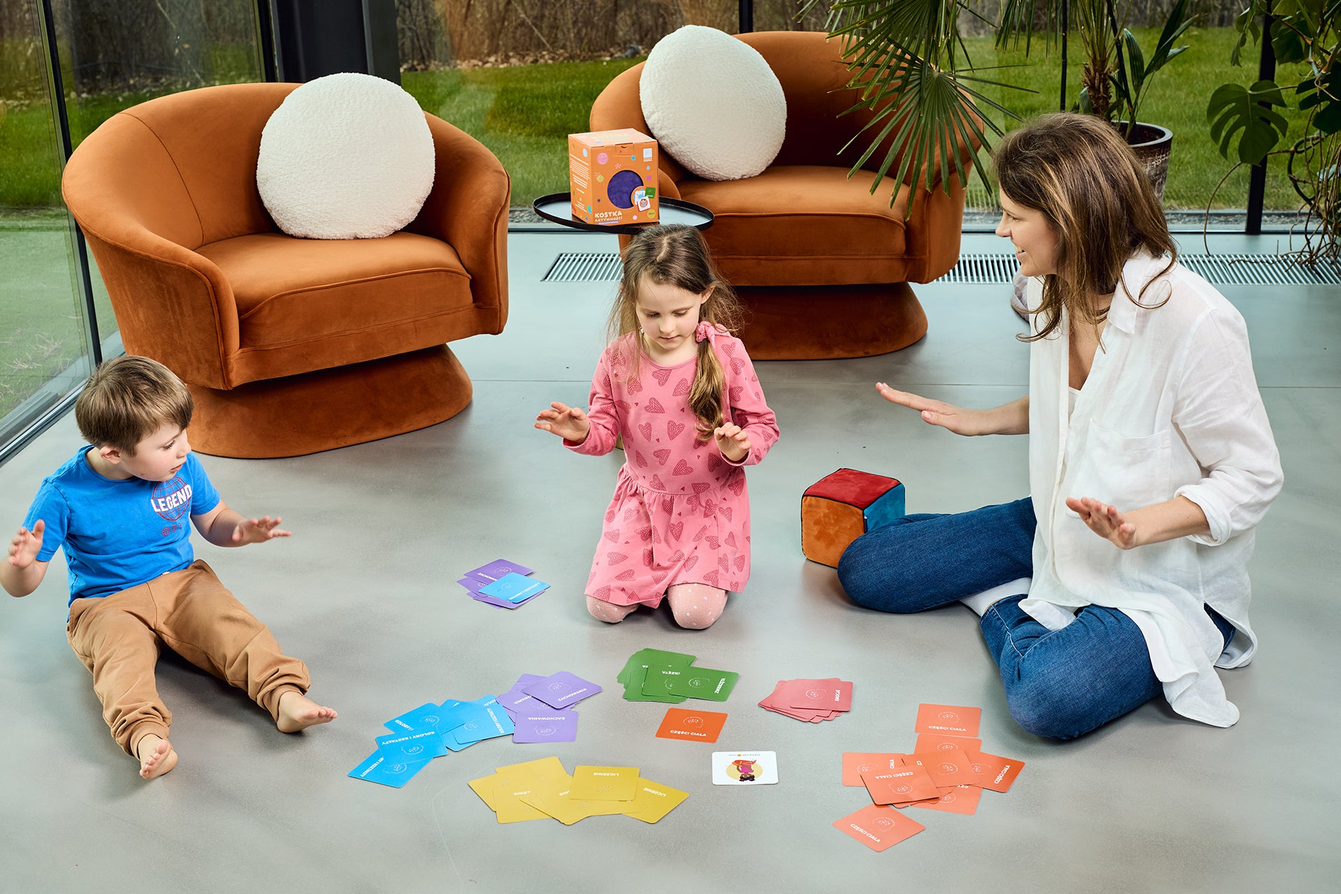 Na zdjęciu widocznych dwoje dzieci - chłopiec i dziewczynka - oraz kobieta. Wszyscy siedzą na podłodze i grają w grę "Kostka aktywności - nauka przez zabawę"